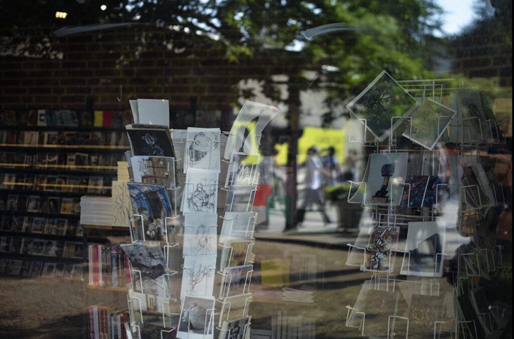 Konsthallstorget speglar sig i bokhandelns fönster. På insidan skymtar vykort .